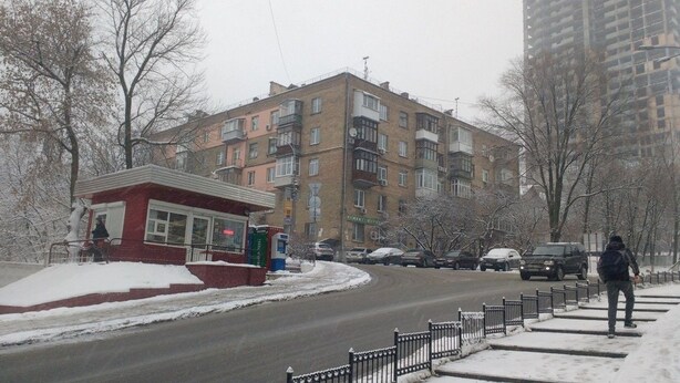 A multi-story building with a red kiosk on the sidewalk, a snow-covered street, and parked cars. - Yourhostel Pechersk (Kyiv)