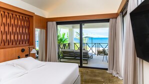 A hotel room with a bed, a TV, a balcony with a view of the ocean, and a seating area with a table and chairs.