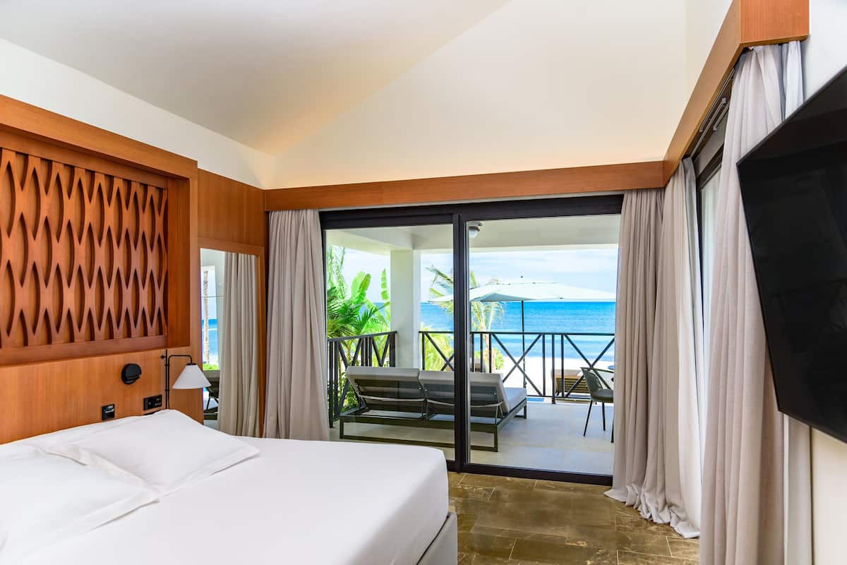 Habitación de hotel con cama, televisión, balcón con vista al océano y zona de estar con mesa y sillas.