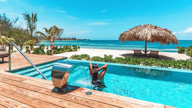 A pool area with a view of the ocean, a thatched umbrella, and lounge chairs.