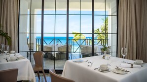 A dining area with tables set for a meal, large windows offering a view of the ocean and palm trees. - The Beach Villas at Excellence Oyster Bay - Adults Only All Inclusive (Falmouth)