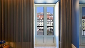 A room with a view of buildings through a glass door, curtains, and a lamp on a side table. - My Story Hotel Figueira (Lisbon)