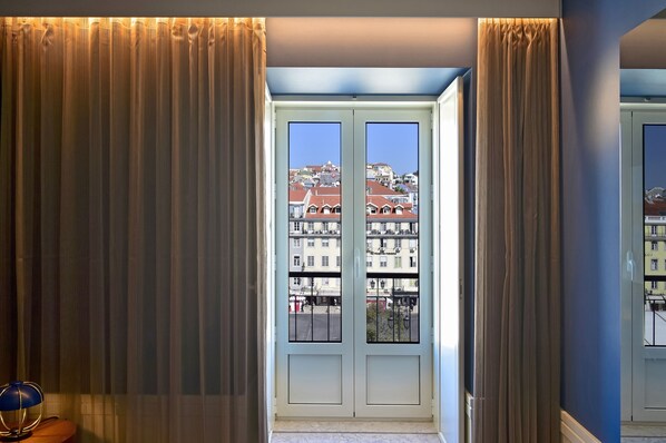 A room with a view of buildings through a glass door, curtains, and a lamp on a side table.