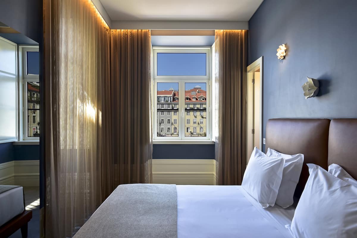 Hotellihuone, jossa on suuri sänky, ikkuna verhoilla, näkymä rakennuksiin ja seinään kiinnitetty valaisin.