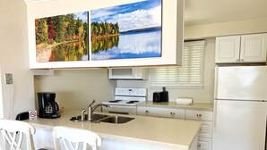 Una cocina moderna con una isla central, armarios blancos, microondas, estufa y refrigerador. Hay dos taburetes de bar y una cafetera sobre la isla.