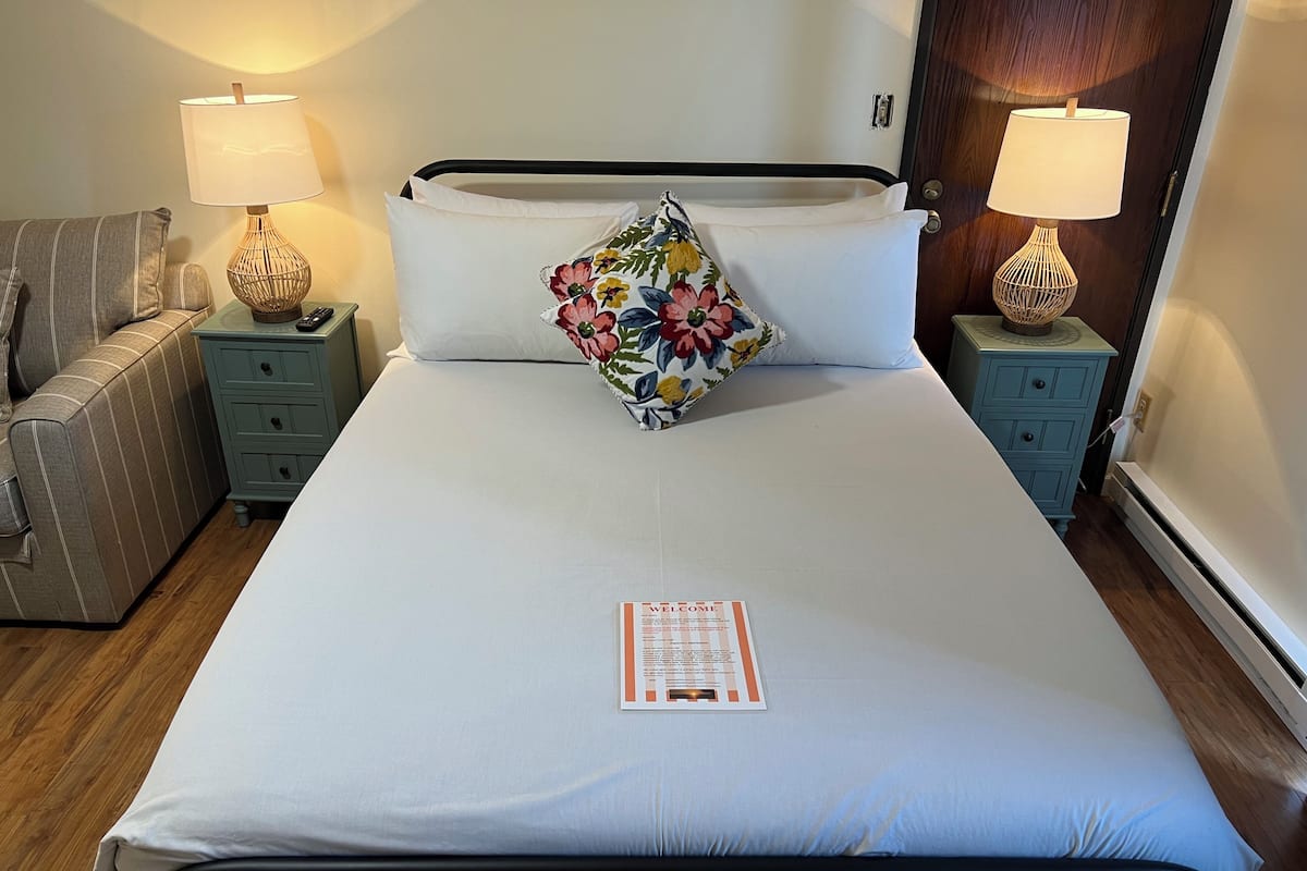 Un lit bien fait, agrémenté d’un coussin à motifs floraux, de deux lampes de chevet, d’une petite table avec une télécommande et d’une carte de bienvenue posée sur le lit.