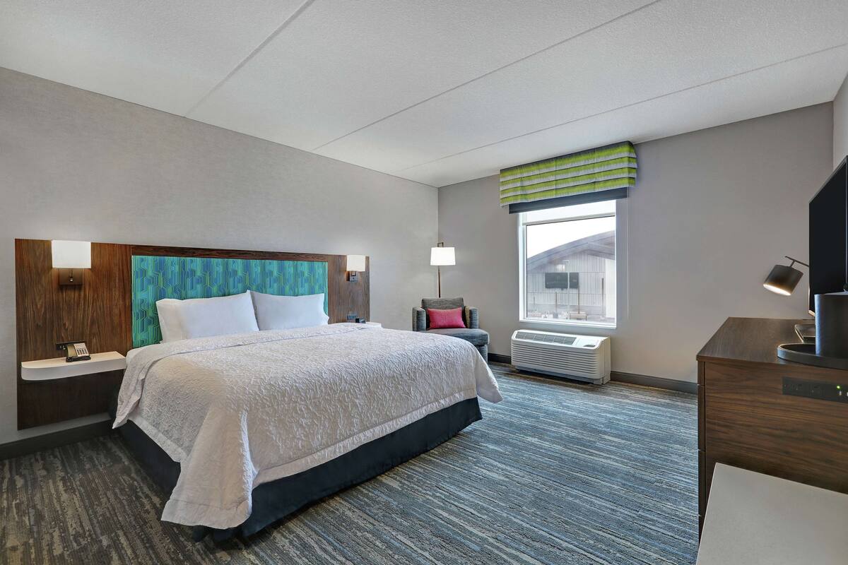 A hotel room with a bed, bedside table, lamp, air conditioning unit, and a window with a view.