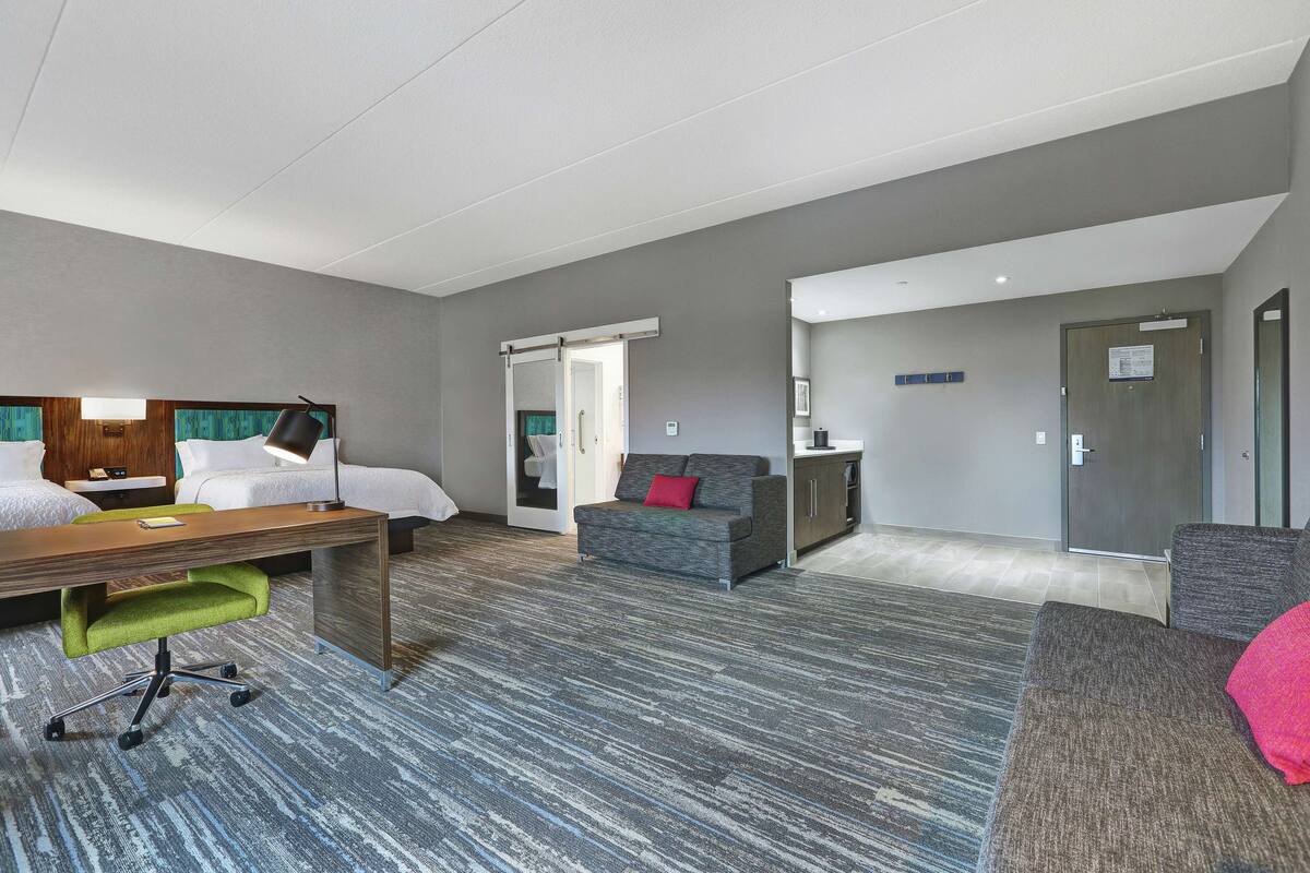 A hotel room with a bed, desk, green office chair, grey sofa, and a small kitchenette.