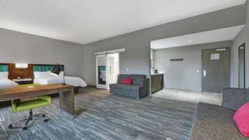 Ein Hotelzimmer mit Bett, Schreibtisch, grünem Bürostuhl, grauem Sofa und einer kleinen Küchenzeile.