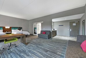 A hotel room with a bed, desk, green office chair, grey sofa, and a small kitchenette.