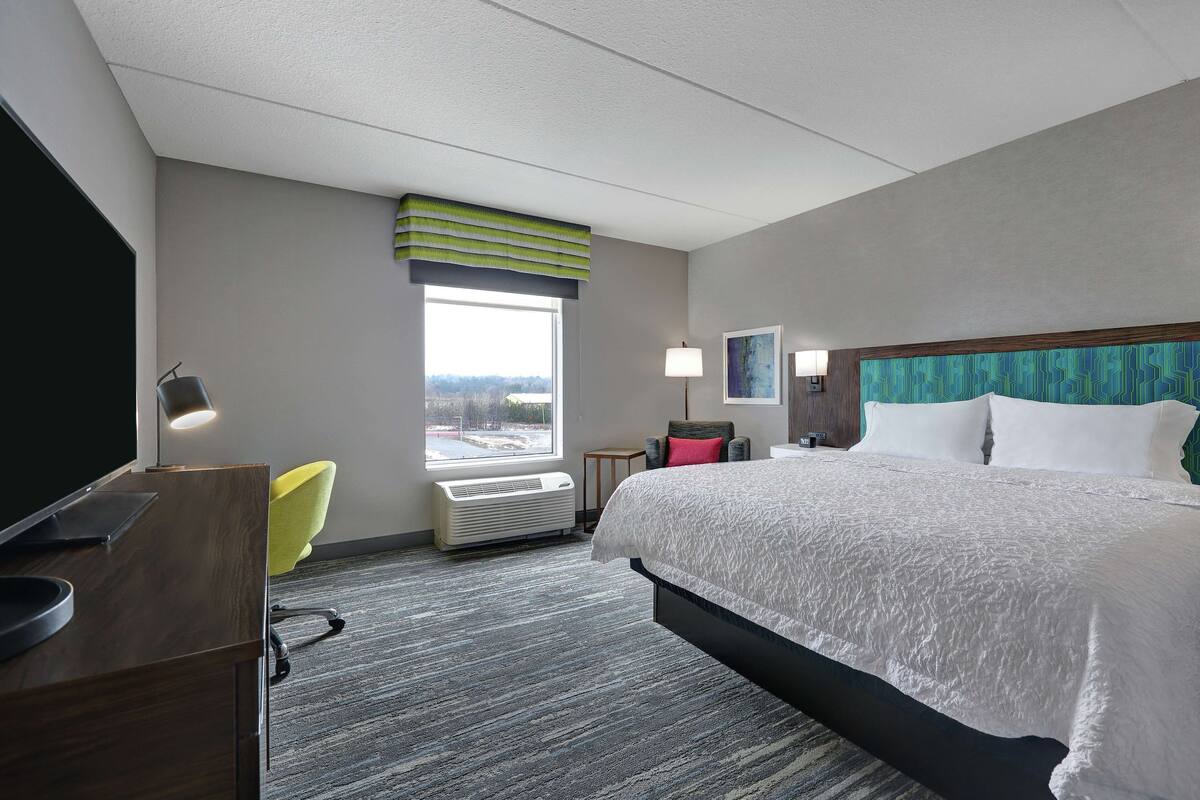 A hotel room with a bed, desk, chair, window, and air conditioning unit.