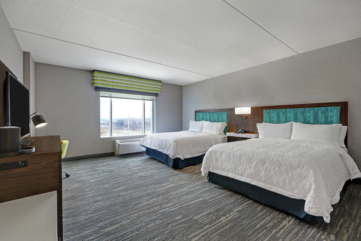 A hotel room with two beds, a desk with a lamp, a window with blinds, and a view of the city.