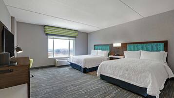 A hotel room with two beds, a desk with a lamp, a window with blinds, and a view of the city.