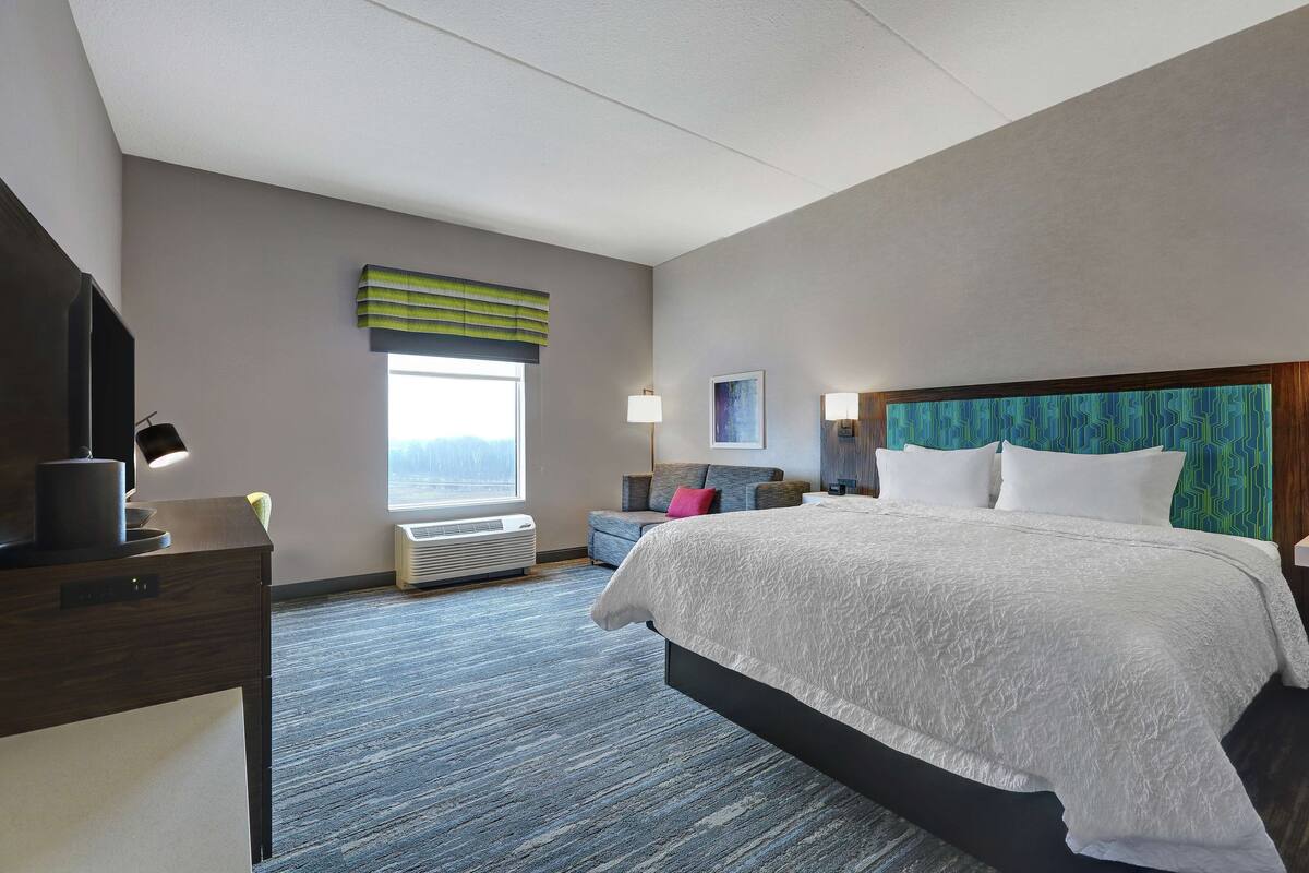 A hotel room with a large bed, a desk with a lamp, a chair, a sofa, a window with a view, and a wall-mounted air conditioner.