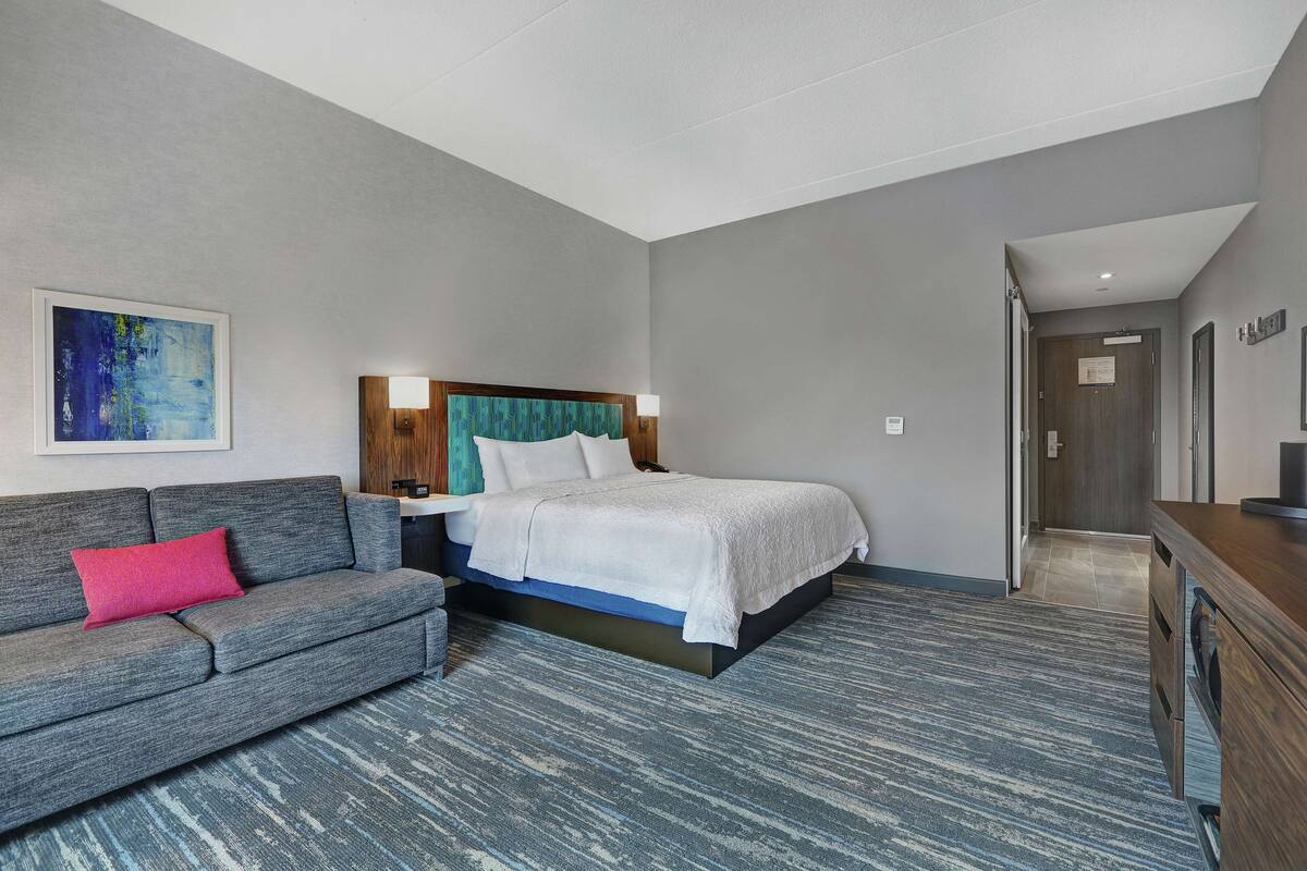 A hotel room with a bed, a gray sofa with a pink pillow, a blue and white patterned carpet, and a wooden headboard.