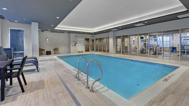 An indoor swimming pool with clear lane markings, a seating area with chairs, and a glass wall offering a view of the gym area. - Hampton Inn by Hilton Peterborough (Peterborough)
