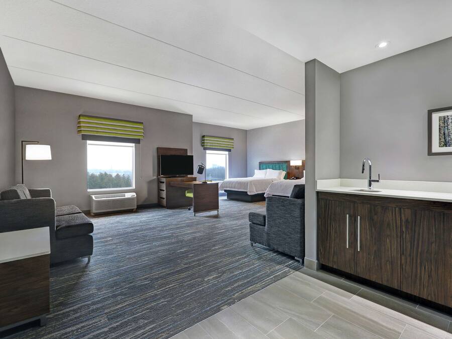 Ein modernes Hotelzimmer mit grauem Teppich, einer grauen Couch, einem Bett mit weißer Bettwäsche, einem Schreibtisch mit Computer und einem Badezimmer mit Waschbecken und Spiegel.