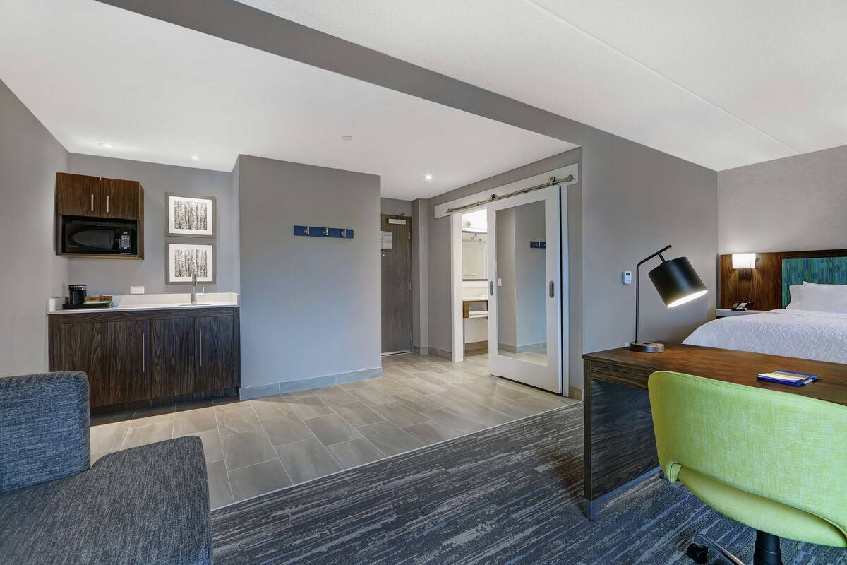 A modern hotel room with a bed, desk, chair, kitchenette, and a bathroom visible through an open door.
