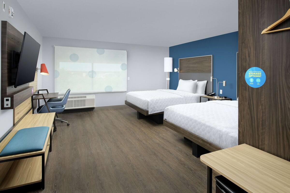 Ein Hotelzimmer mit zwei Betten, einem Schreibtisch, einem Stuhl, einem Fernseher und einem Fenster mit Jalousien.