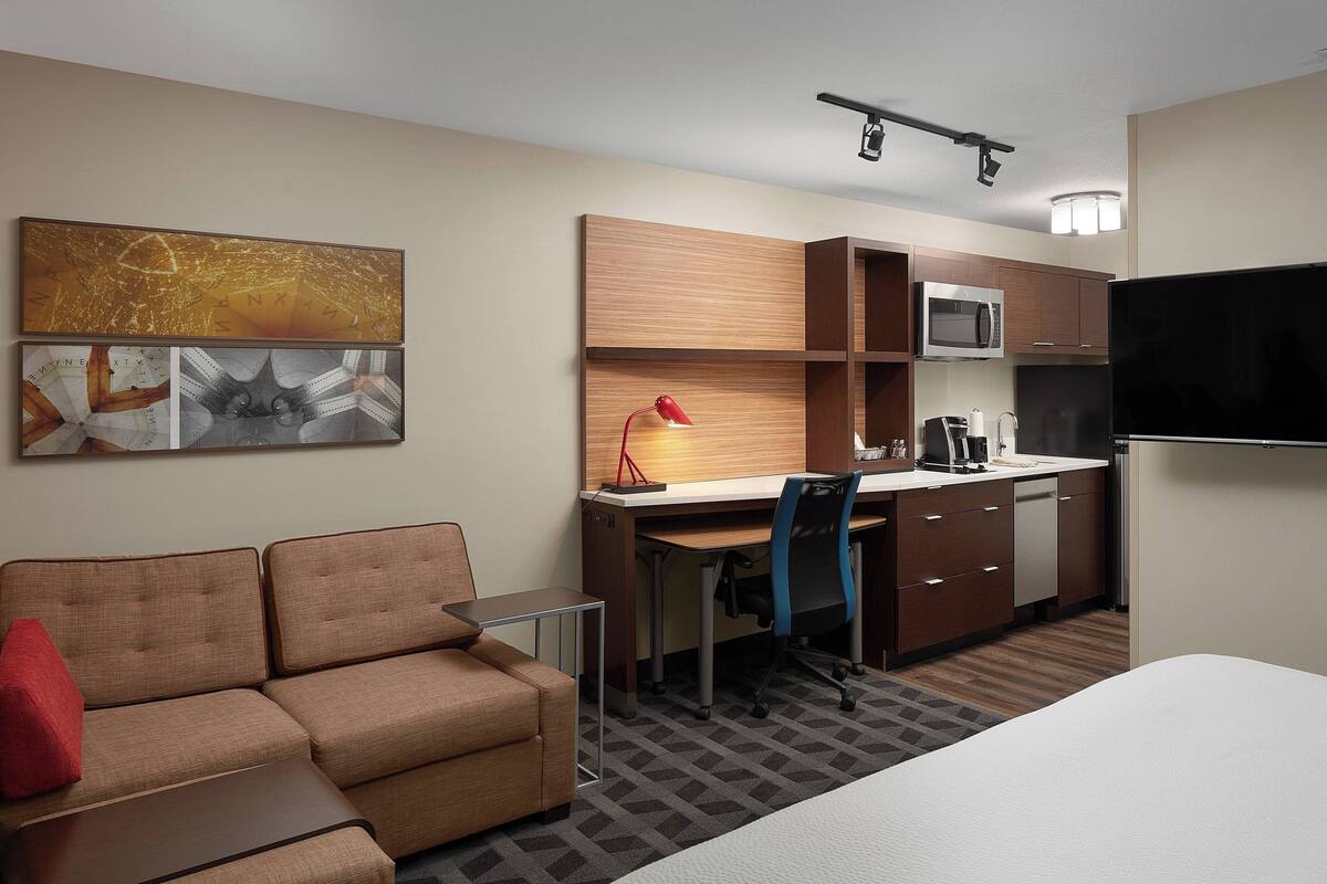 Habitación de hotel con sofá, escritorio y silla, microondas, refrigerador y televisor de pantalla plana.