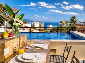 A poolside dining area with a view of the sea, a table set with plates and a glass of orange juice, and a chair.