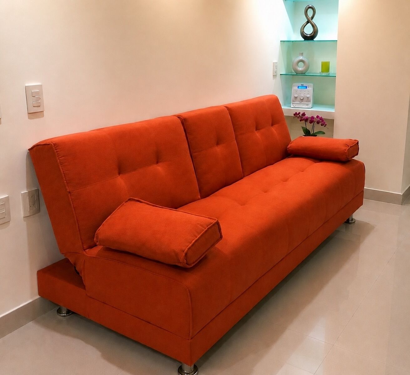 Un canapé orange moderne avec un coussin, placé dans une pièce dont le sol est de couleur claire et où l’on aperçoit, au fond, une petite étagère ornée d’objets décoratifs.