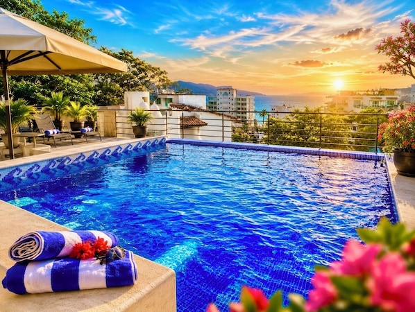 A swimming pool with a tiled edge, lounge chairs, and a view of the sea at sunset.