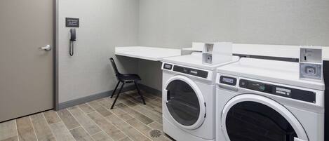 A laundry room with two front-loading washing machines, a chair, a phone, and a sign indicating assistance.