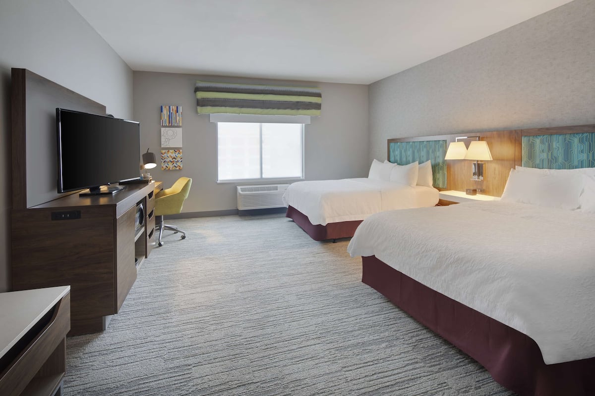 A hotel room with a flat-screen TV, a desk, two beds, a chair, a window, and a wall-mounted lamp.