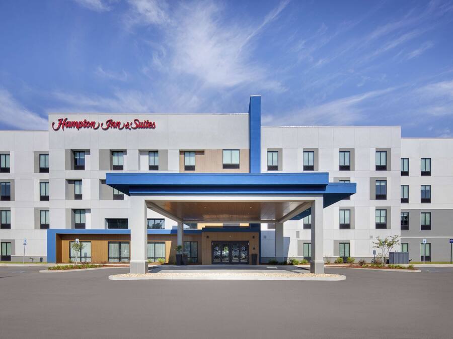 Um hotel Hampton Inn & Suites com design moderno, apresentando uma entrada coberta e um céu azul límpido.
