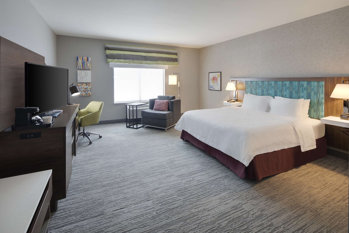 A hotel room with a large bed, a desk with a computer, a chair, a TV, and a window with a view.