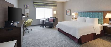 A hotel room with a large bed, a desk with a computer, a chair, a TV, and a window with a view.