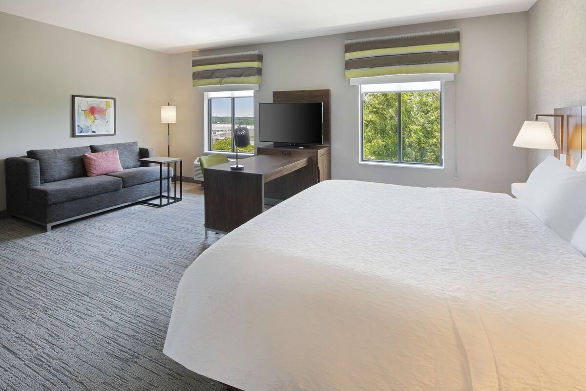 A hotel room with a large bed, a desk with a flat-screen TV, a sofa, and two windows with curtains.