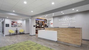 A reception area with a wooden reception desk, a 'Welcome to the Magnolia State' sign, a bookshelf with various items, and two yellow chairs. - Hampton Inn & Suites D'Iberville Biloxi (D'Iberville)