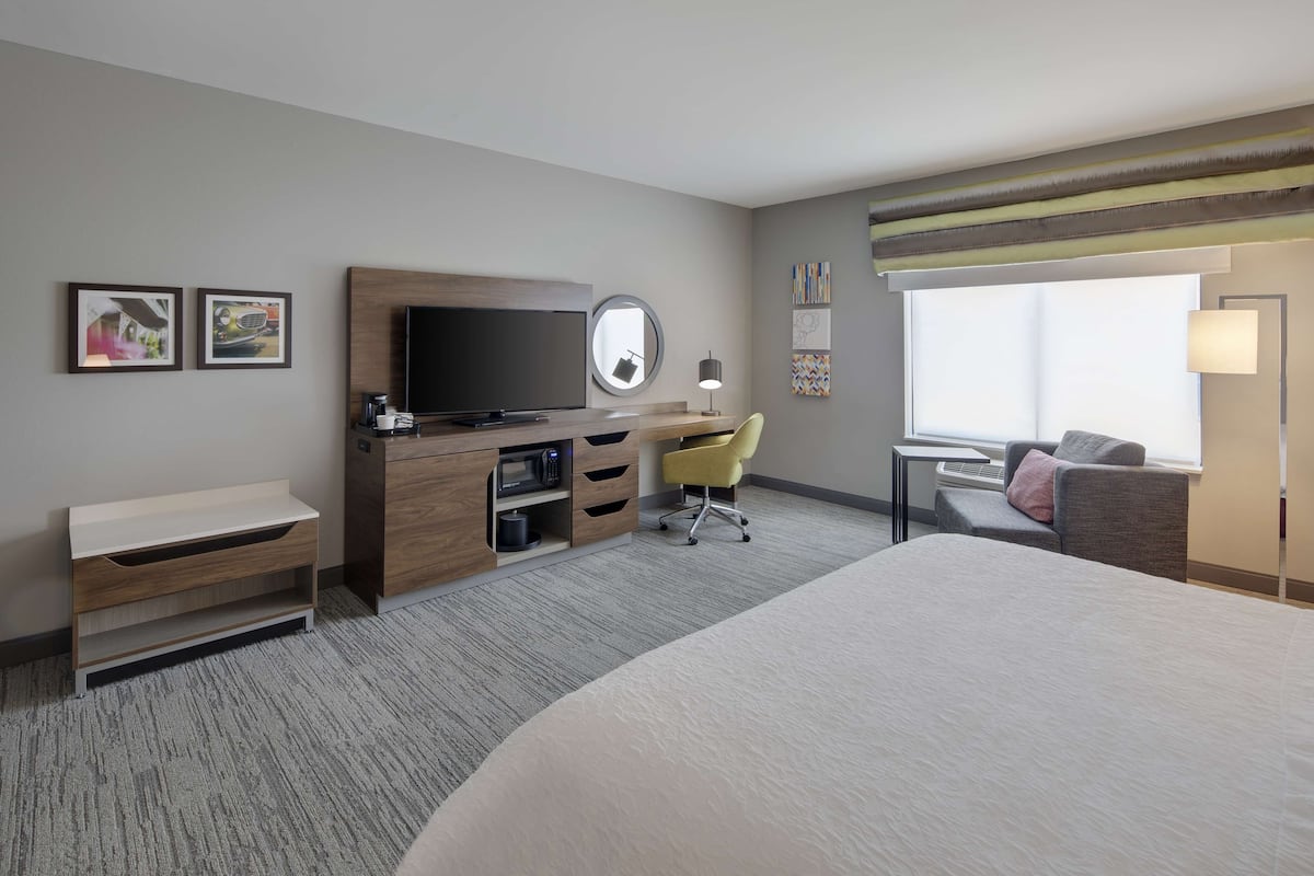 A hotel room with a bed, a desk with a TV, a chair, a small table, and a window.