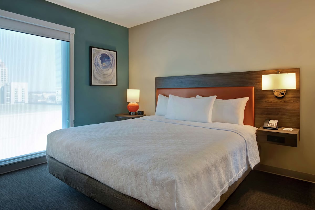 Une chambre d’hôtel avec un grand lit, une table de chevet avec une lampe, un téléphone fixé au mur et une fenêtre donnant sur des immeubles.