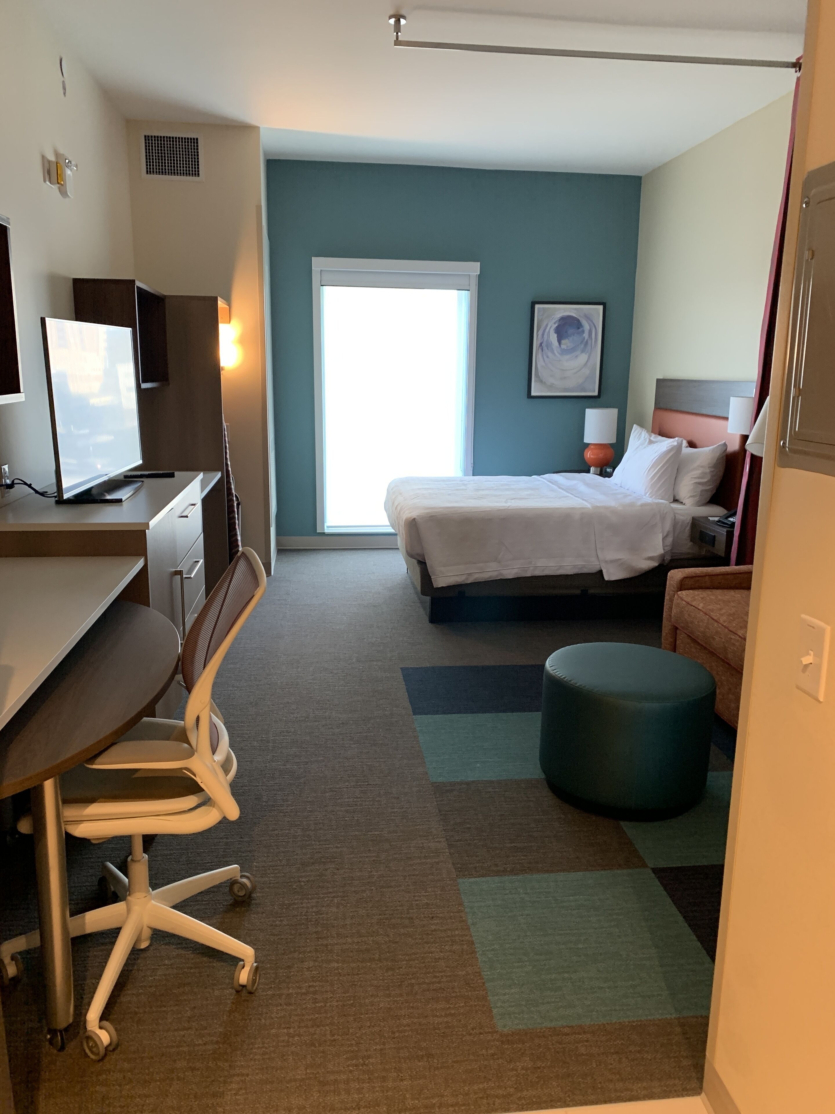 A hotel room with a desk, chair, bed, and ottoman.