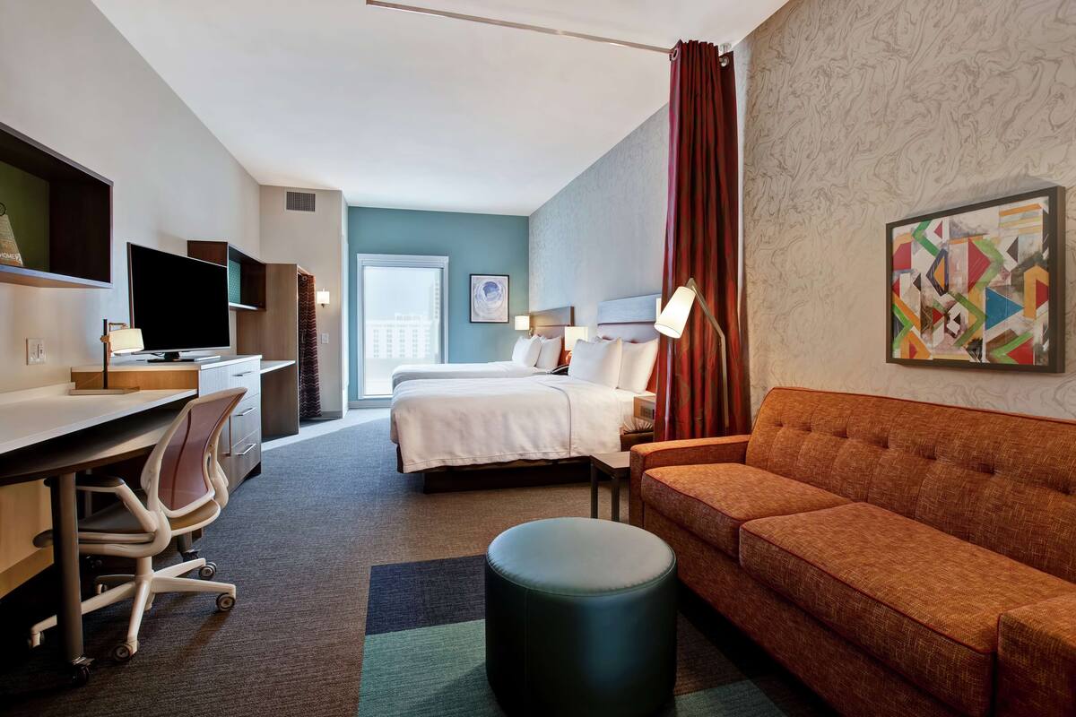 Une chambre d’hôtel équipée d’un lit, d’un bureau, d’une chaise, d’un pouf, d’un canapé, d’une télévision et d’une fenêtre donnant sur des immeubles.