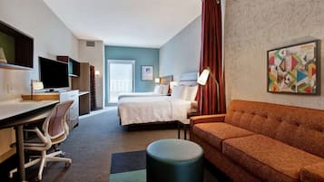 A hotel room with a bed, a desk, a chair, an ottoman, a sofa, a TV, and a window with a view of buildings.