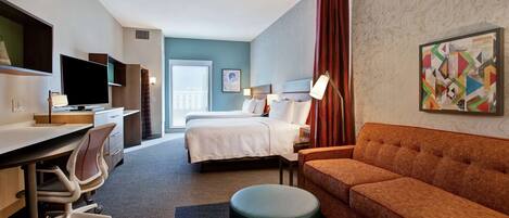 A hotel room with a bed, a desk, a chair, an ottoman, a sofa, a TV, and a window with a view of buildings.