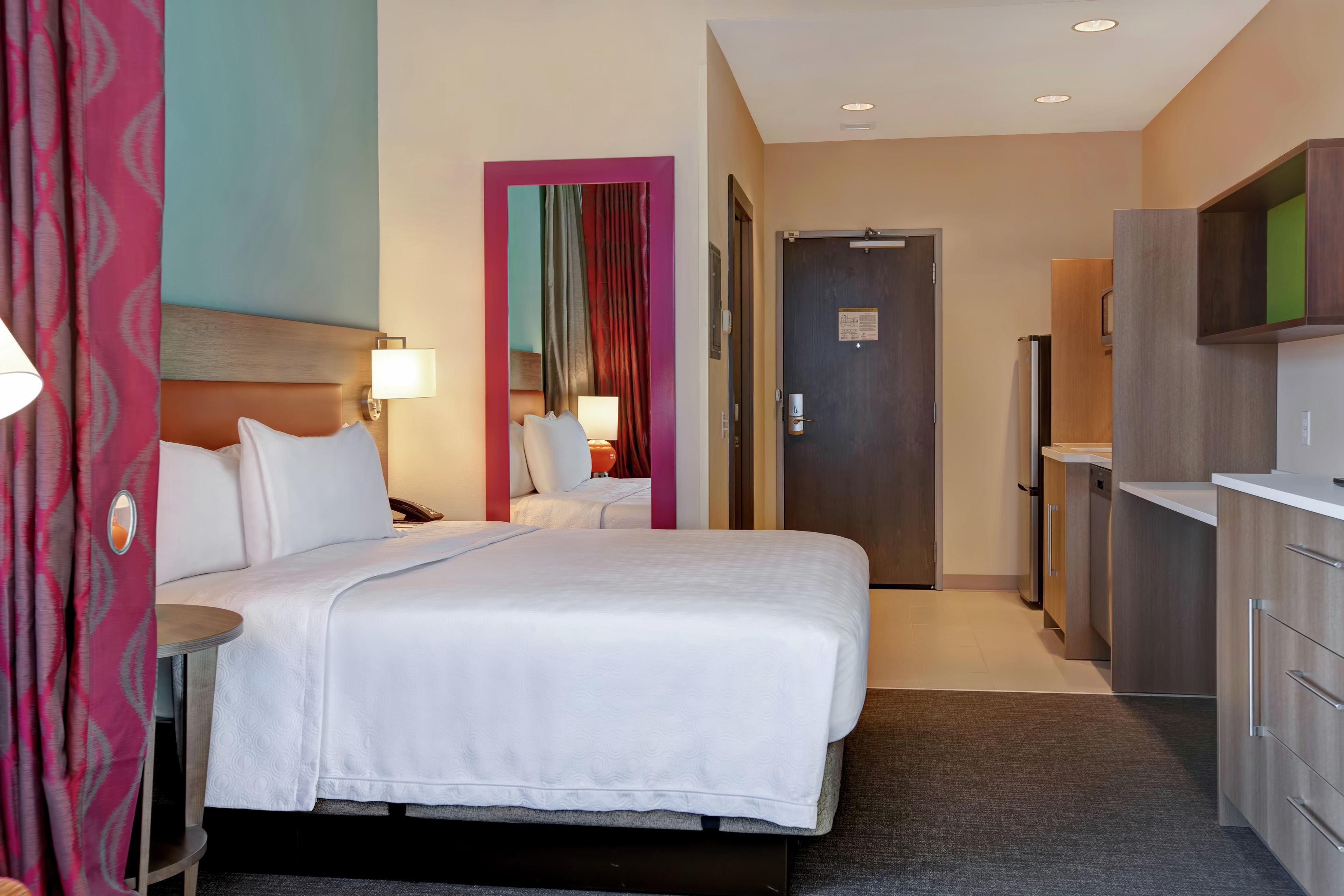 A hotel room with a bed, bedside tables, a desk, a mirror, a door, and a kitchenette.
