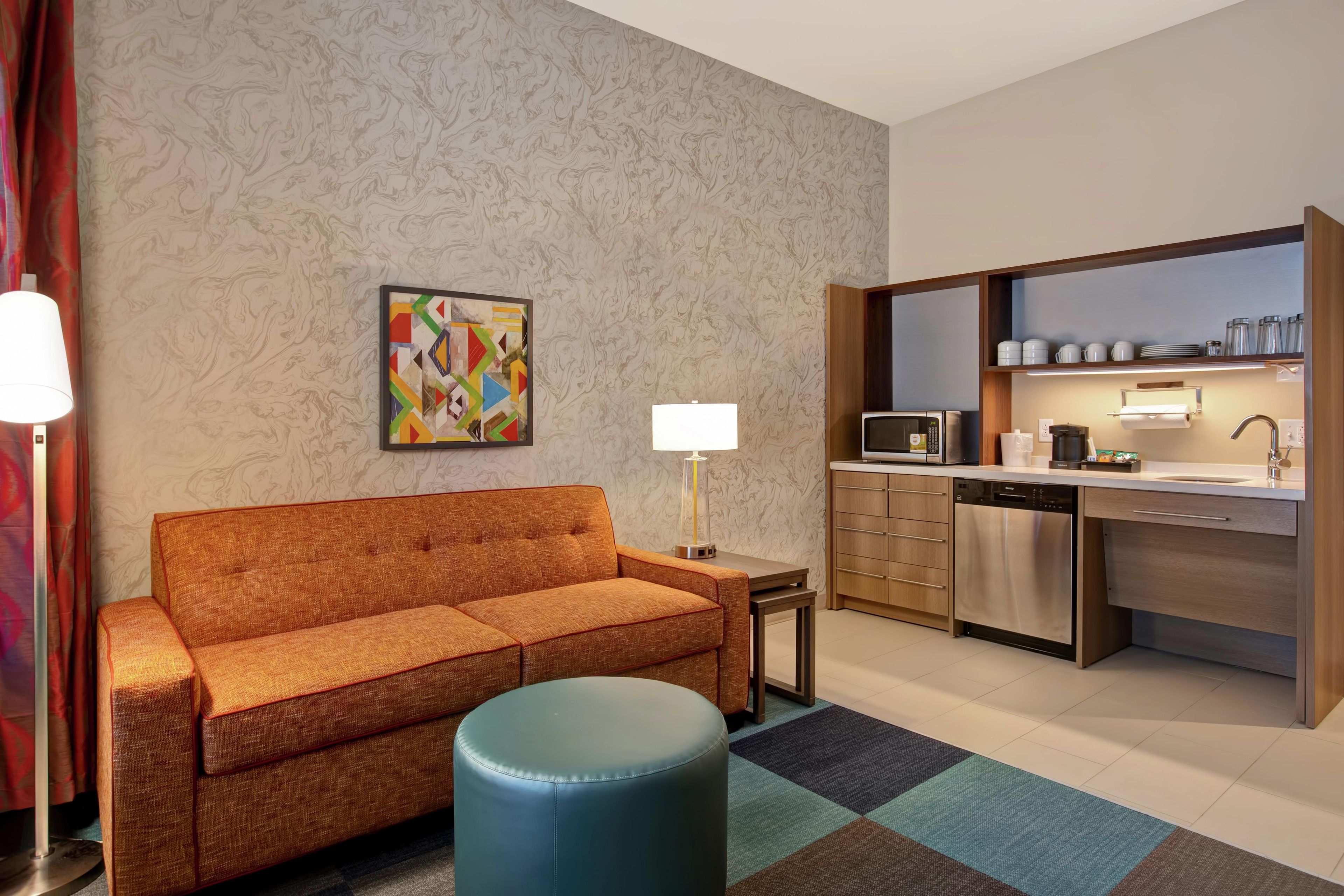 A hotel room with a sofa, a small table, a lamp, a colorful painting, a kitchenette, and a blue ottoman.