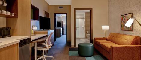 A hotel room with a kitchenette, a sofa, a TV, and a bathroom visible through an open door.