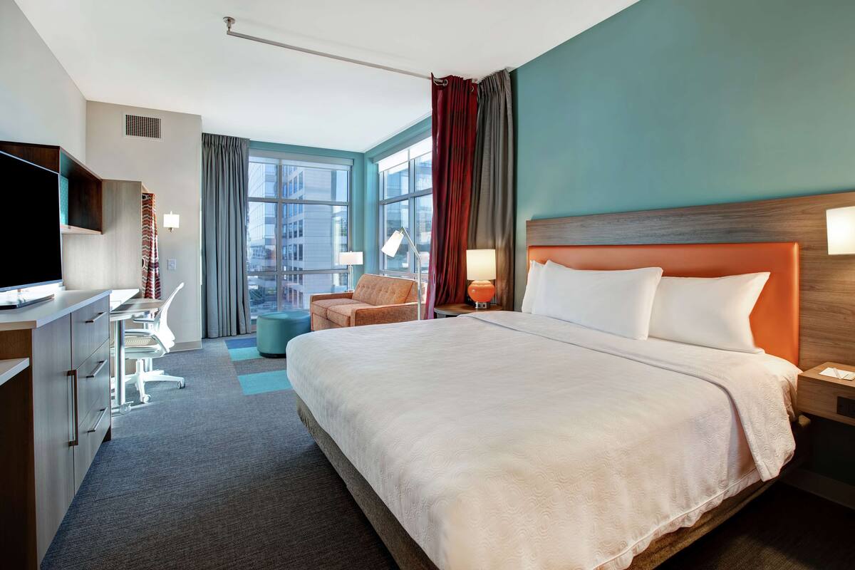 Une chambre d’hôtel avec un grand lit, un bureau avec une chaise, une télévision et une fenêtre avec des rideaux.