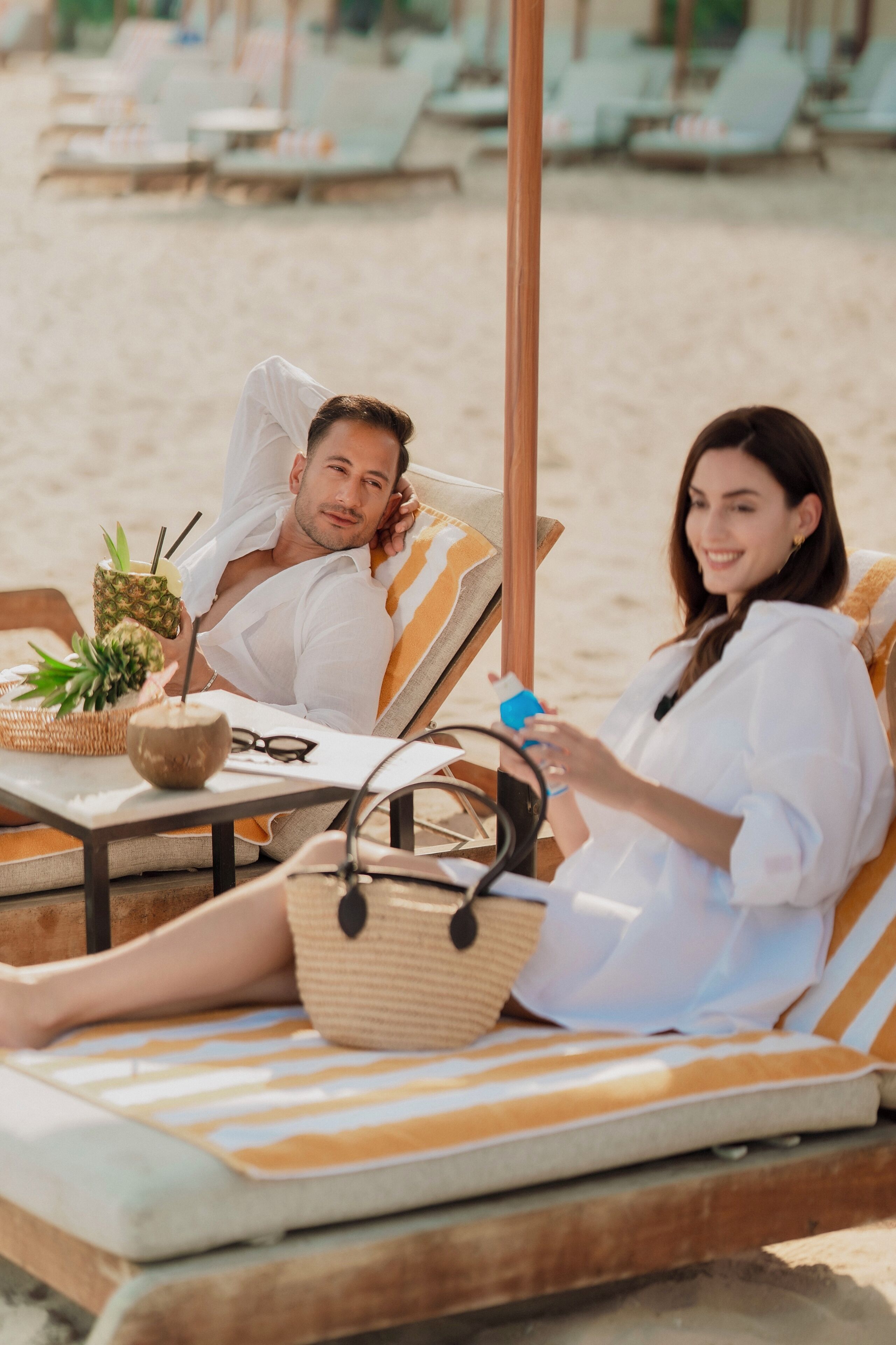 Un área de descanso junto a la playa con cojines a rayas y una mesa con una bebida, una cesta y un coco.