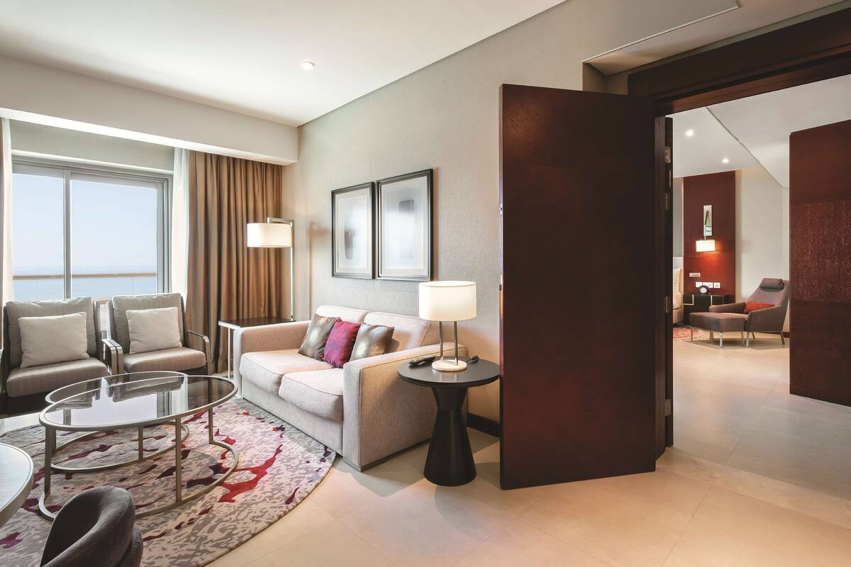 Une chambre d’hôtel moderne, dotée d’un canapé, de fauteuils, d’une table basse et offrant une vue sur la mer grâce à de grandes fenêtres.
