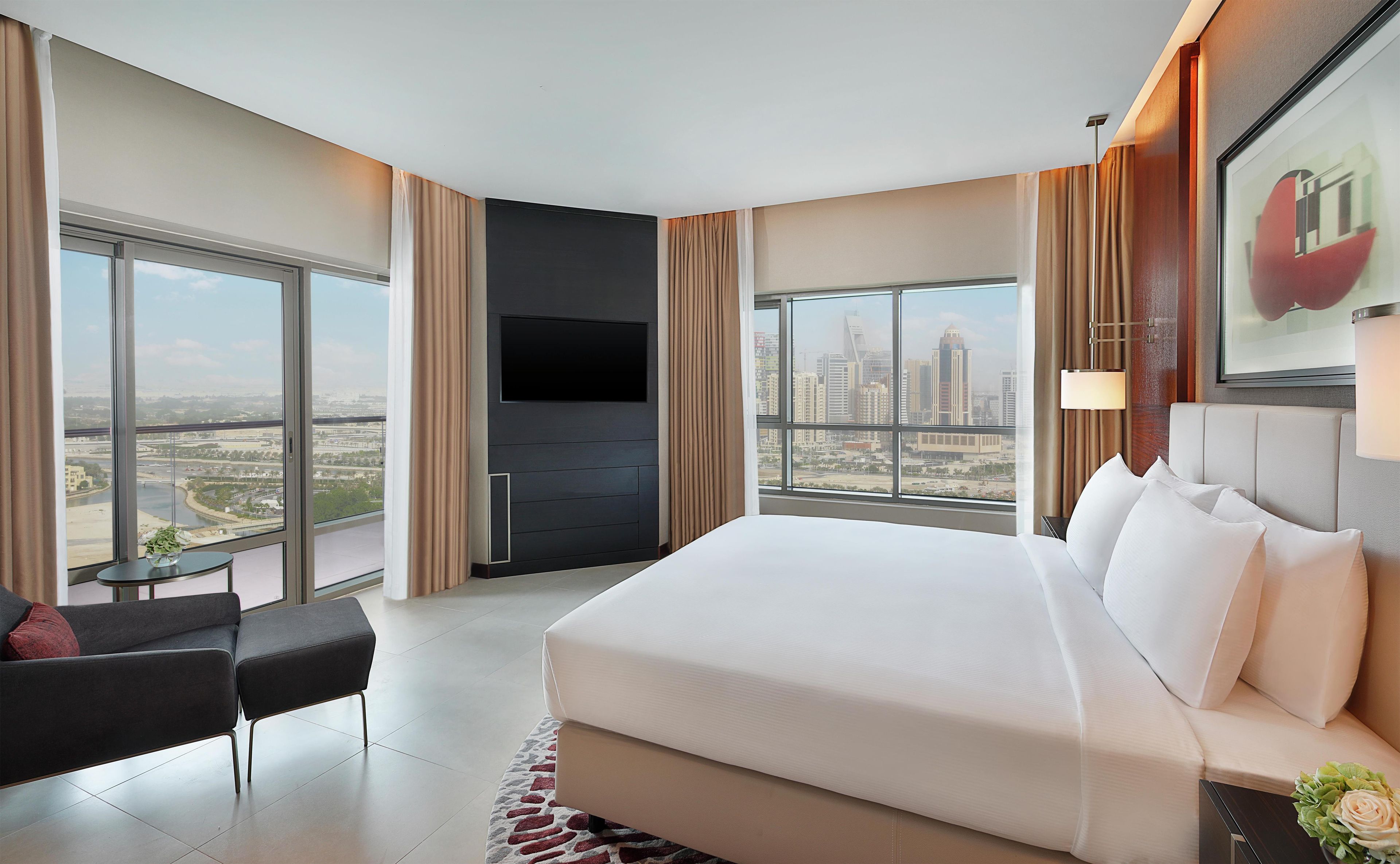 A hotel room with a large bed, a TV mounted on the wall, a desk, and a view of the cityscape.