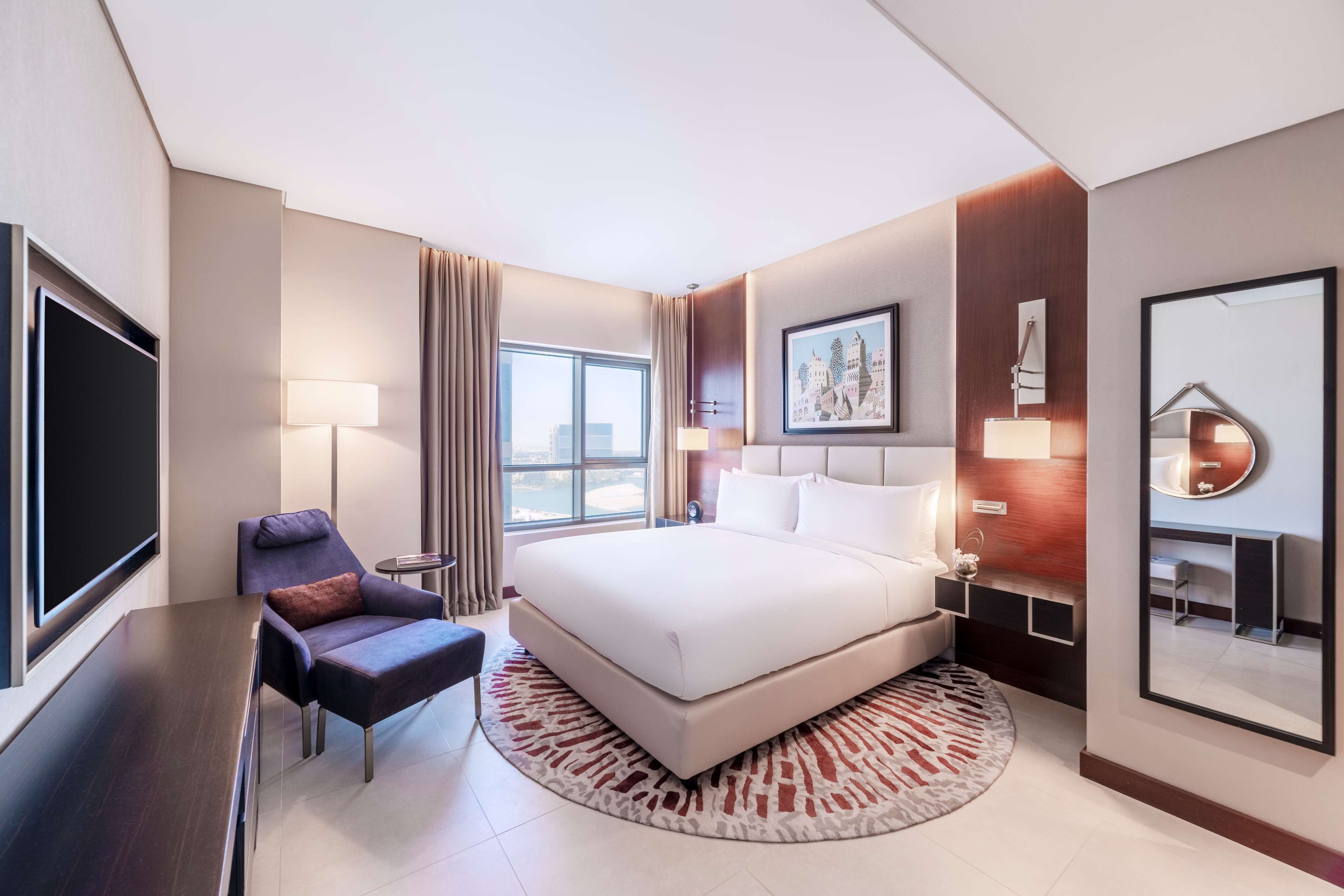 A modern hotel room with a large bed, a desk, a chair, a TV, and a window with curtains.