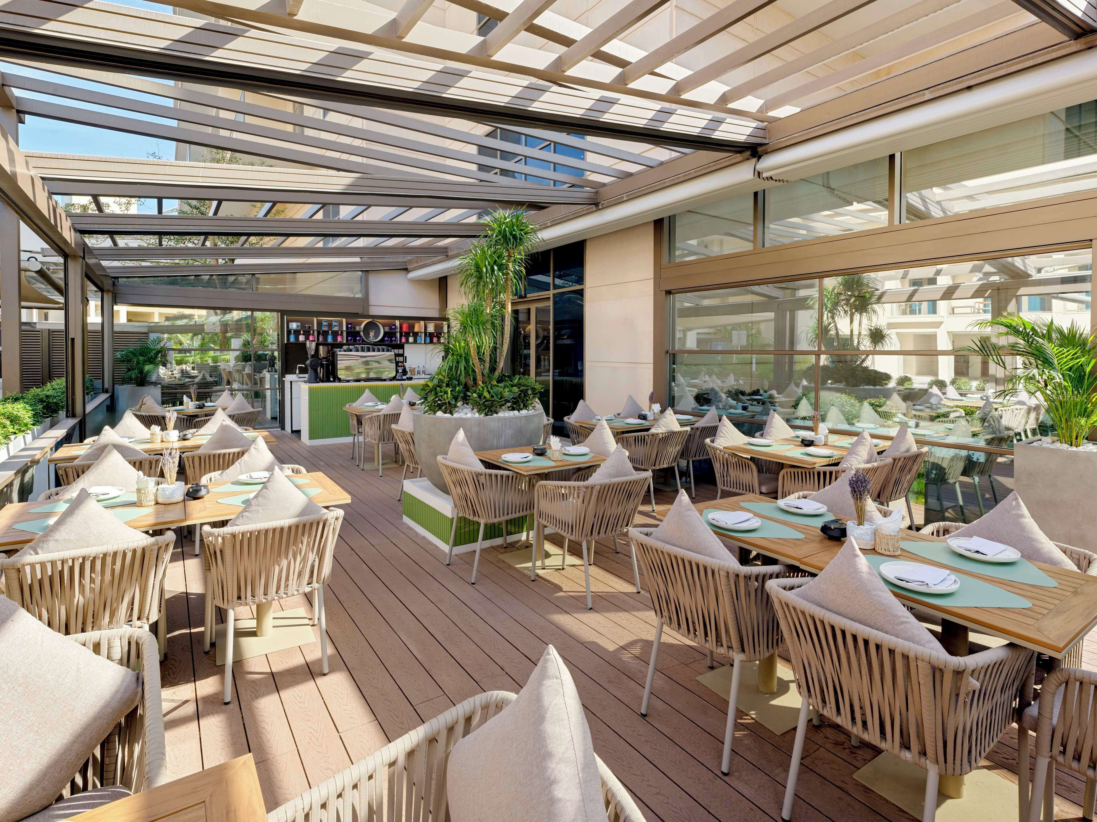 An outdoor café with wooden tables and wicker chairs, a bar area, and a large window offering a view of the surroundings.