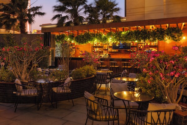 An outdoor bar area with tables and chairs, surrounded by plants and palm trees. - Hilton Doha The Pearl (Doha)